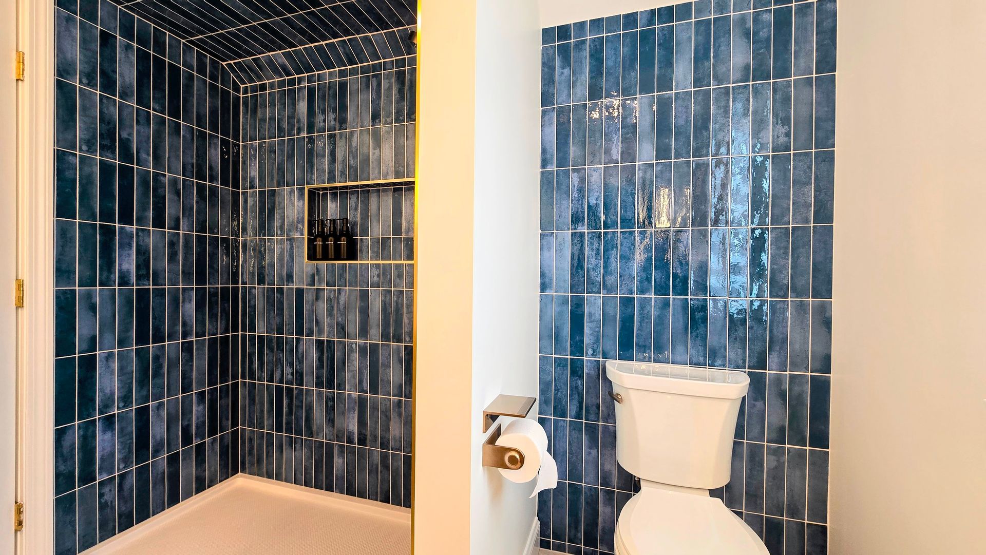 A bathroom with a tiled shower stall and a toilet, featuring deep blue, vertically stacked rectangular tiles.