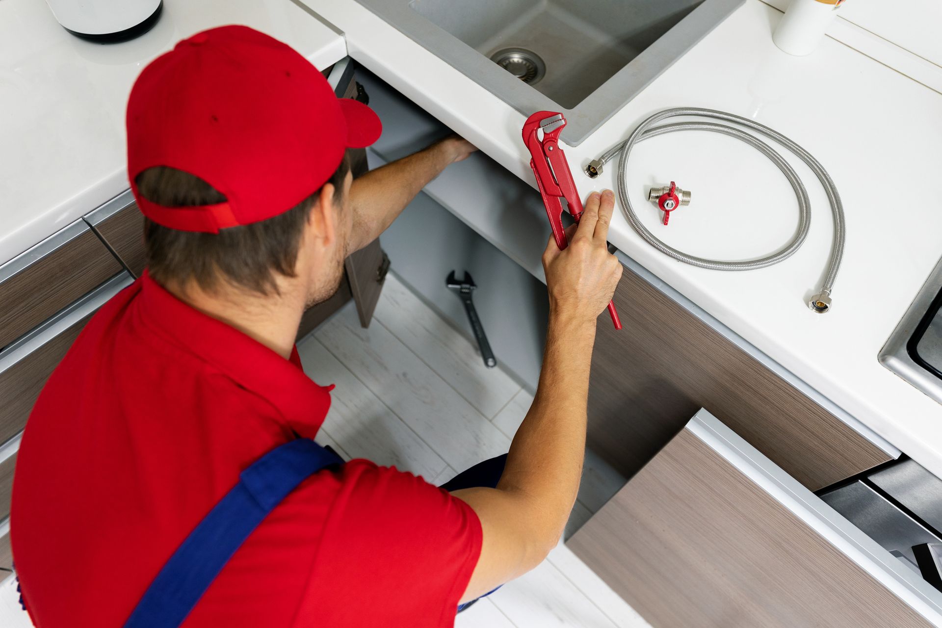 Plumber in red uniform repairing kitchen sink with wrench and hose, top view of plumbing work.