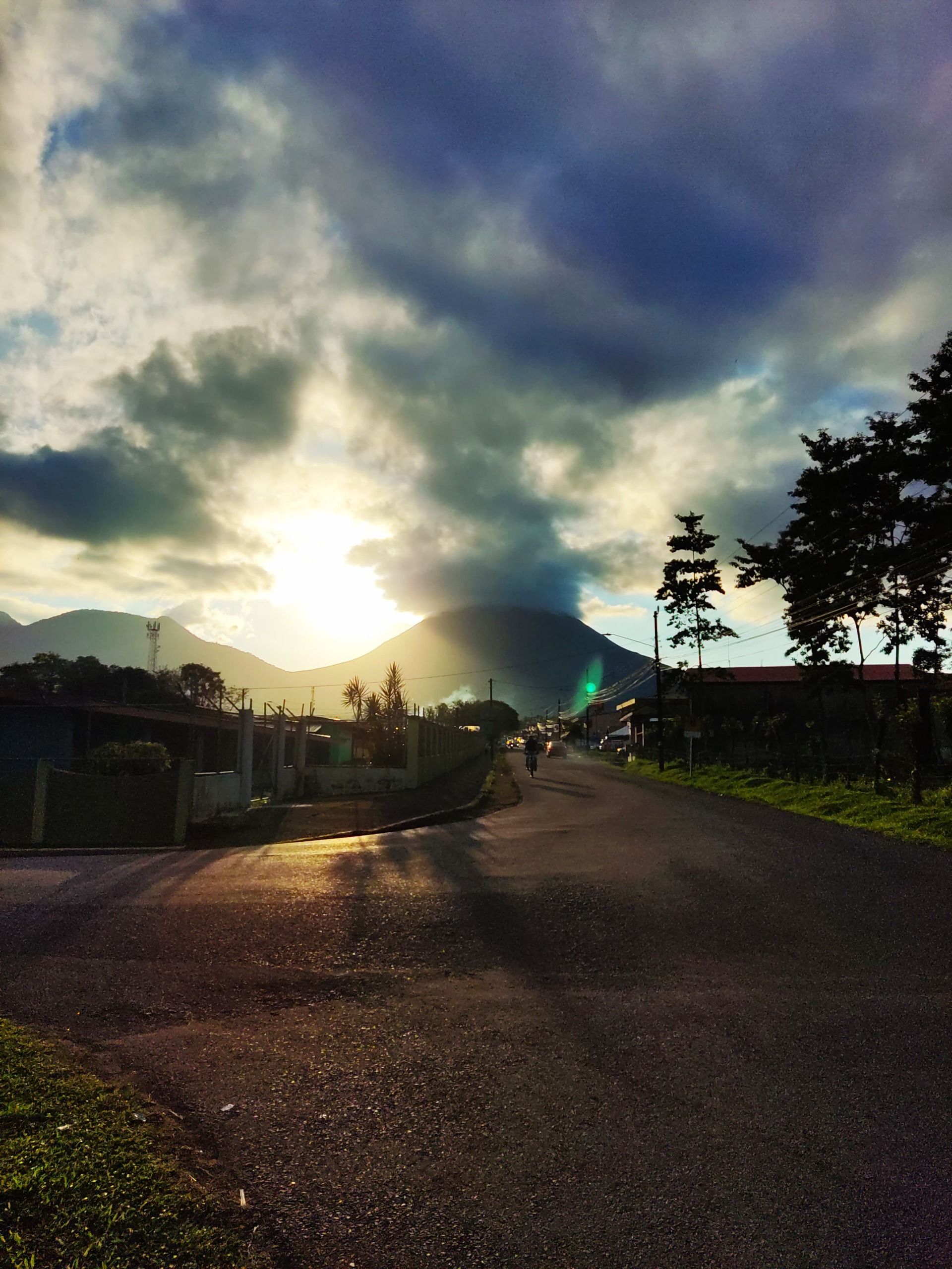 a road with a mountain in the background and the sun shining through the clouds