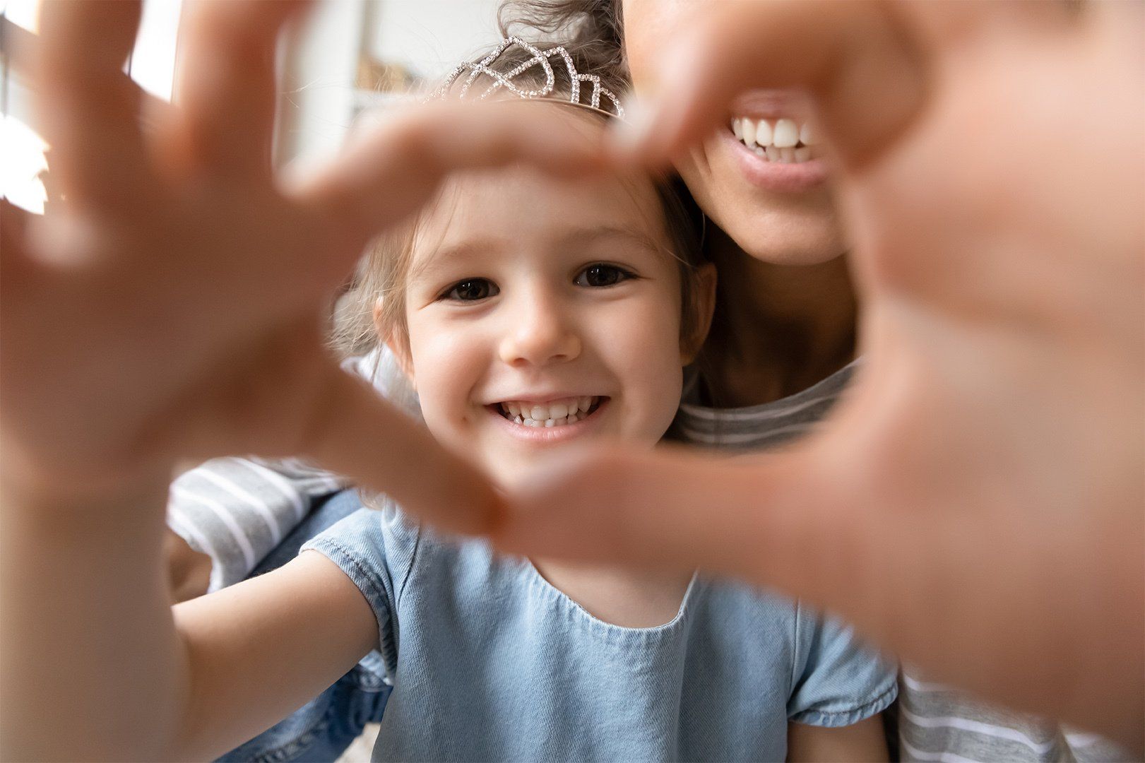 Close up little girl wearing princess diadem and mother showing heart sign with hands