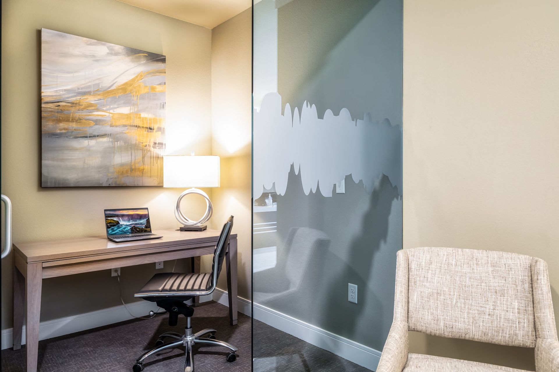 A small office with a desk , chair , laptop and painting on the wall.