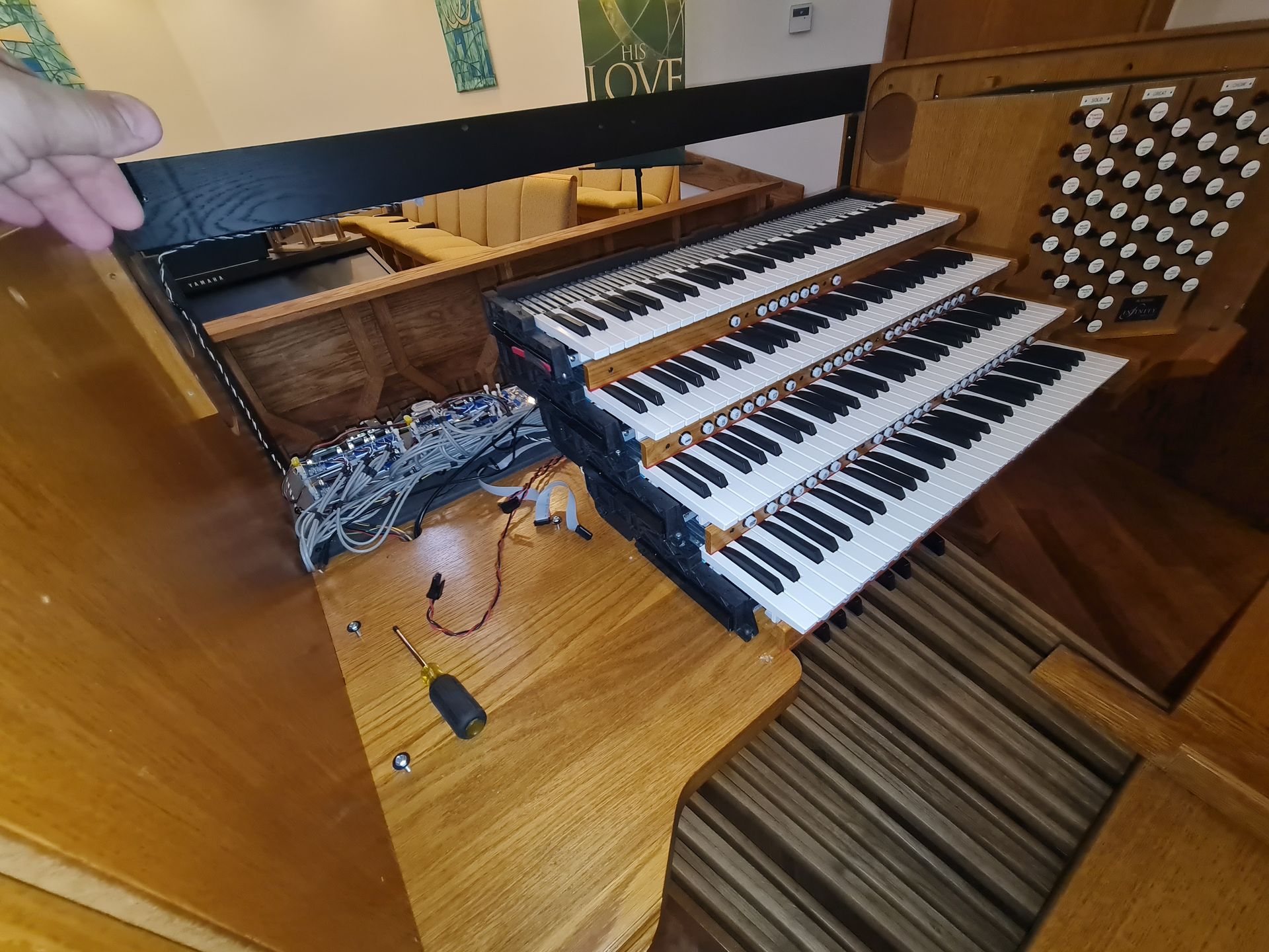 An organ console with three keyboards and pedalboard, surrounded by wooden panels, and a hand.