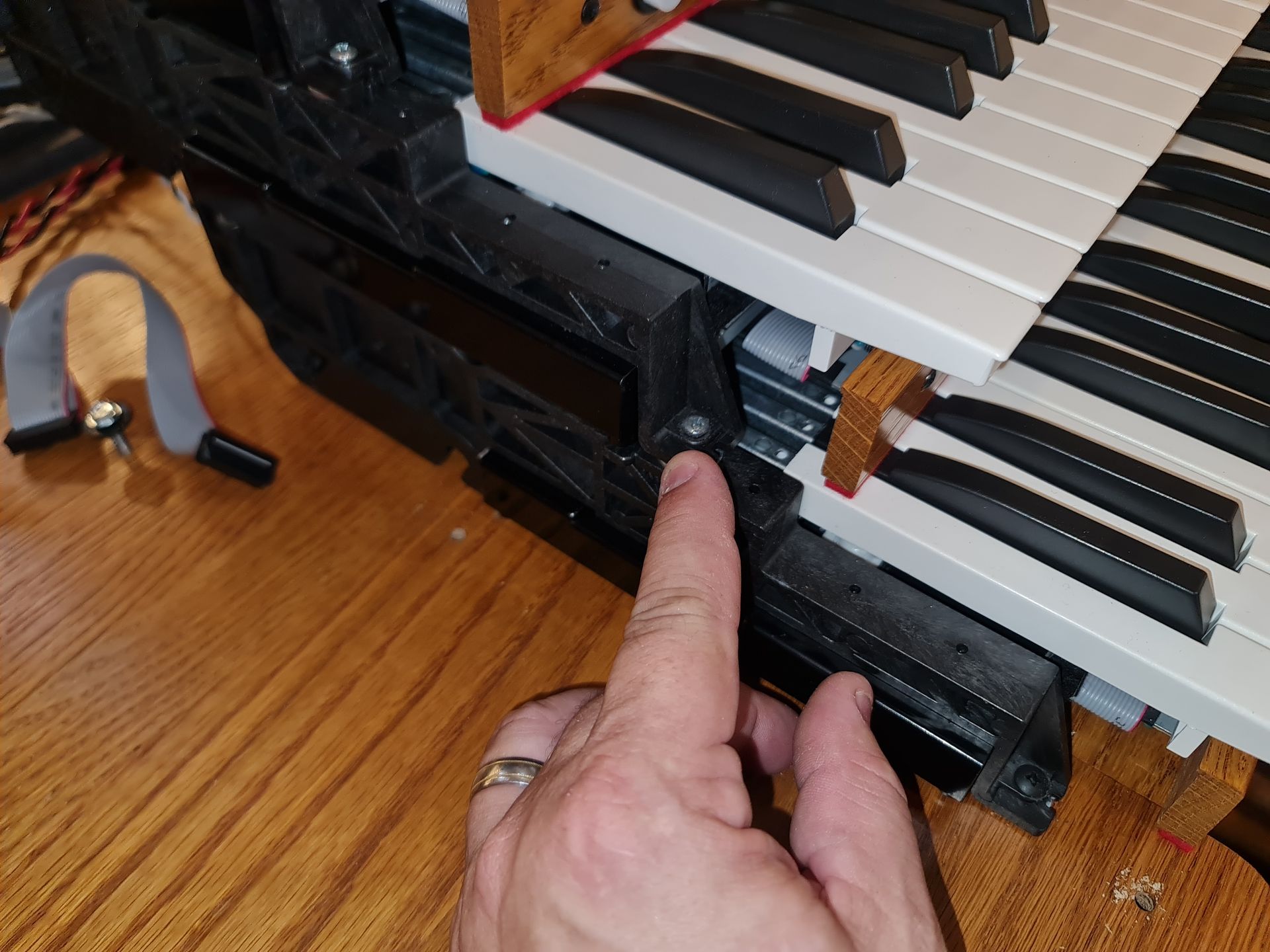 Hand pointing at a screw on a black plastic keyboard frame next to white and black keys.