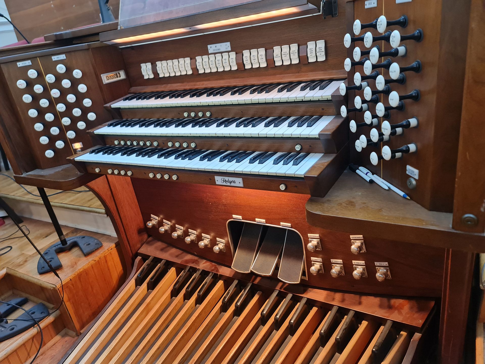 Close-up of a pipe organ with three keyboards, numerous buttons, and foot pedals. Brown wood construction.