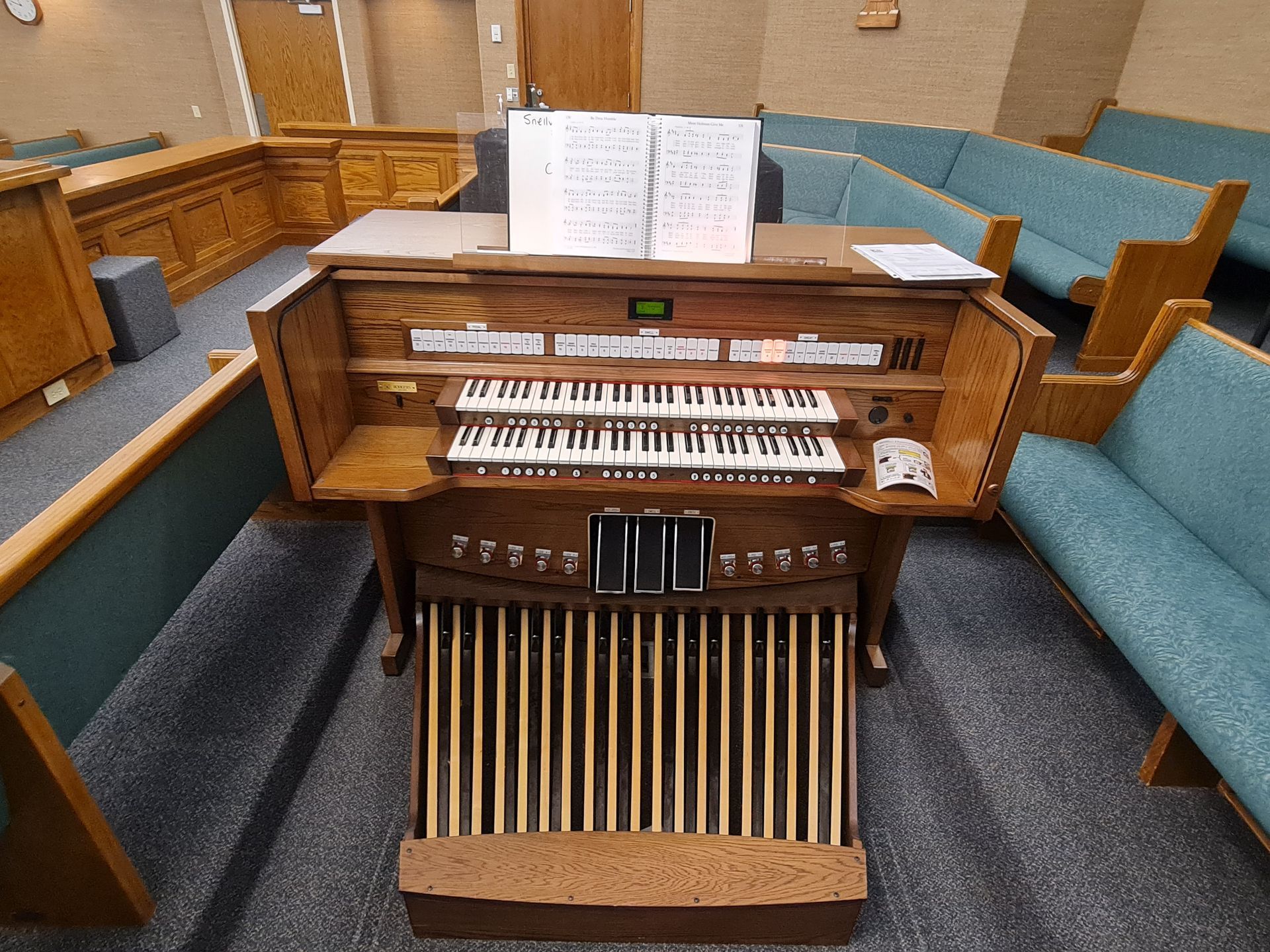 Church organ with keyboards, foot pedals, and sheet music, surrounded by pews in a sanctuary.