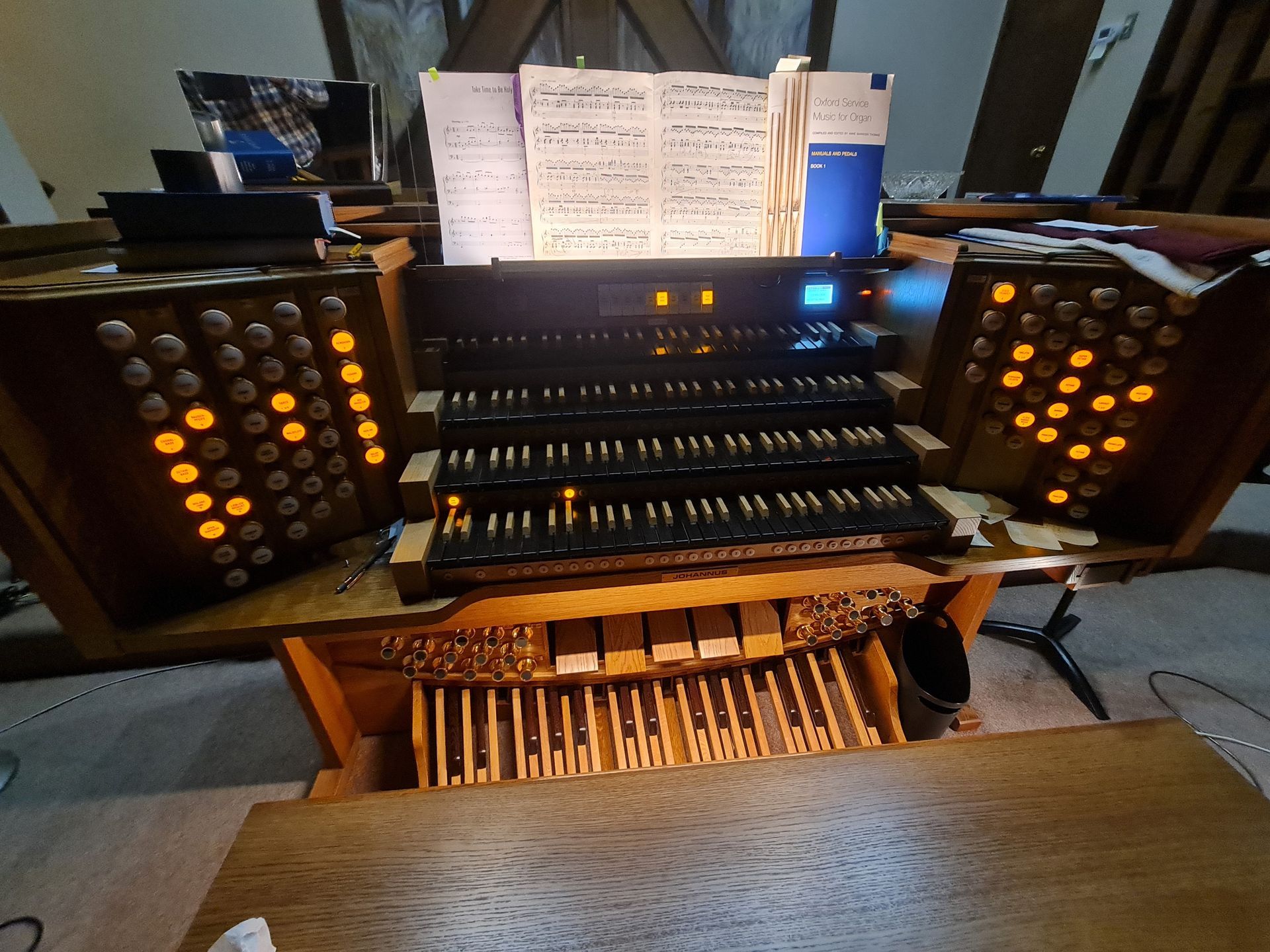 Organ console with multiple keyboards, pedals, and illuminated stop tabs; sheet music on top.