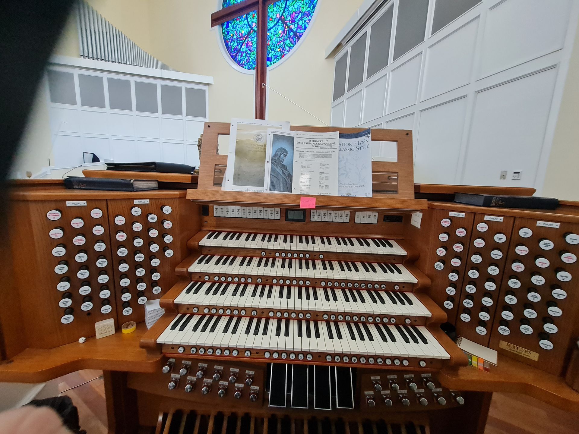 Church organ with multiple keyboards and stops, music on the stand, with a stained glass window in the background.