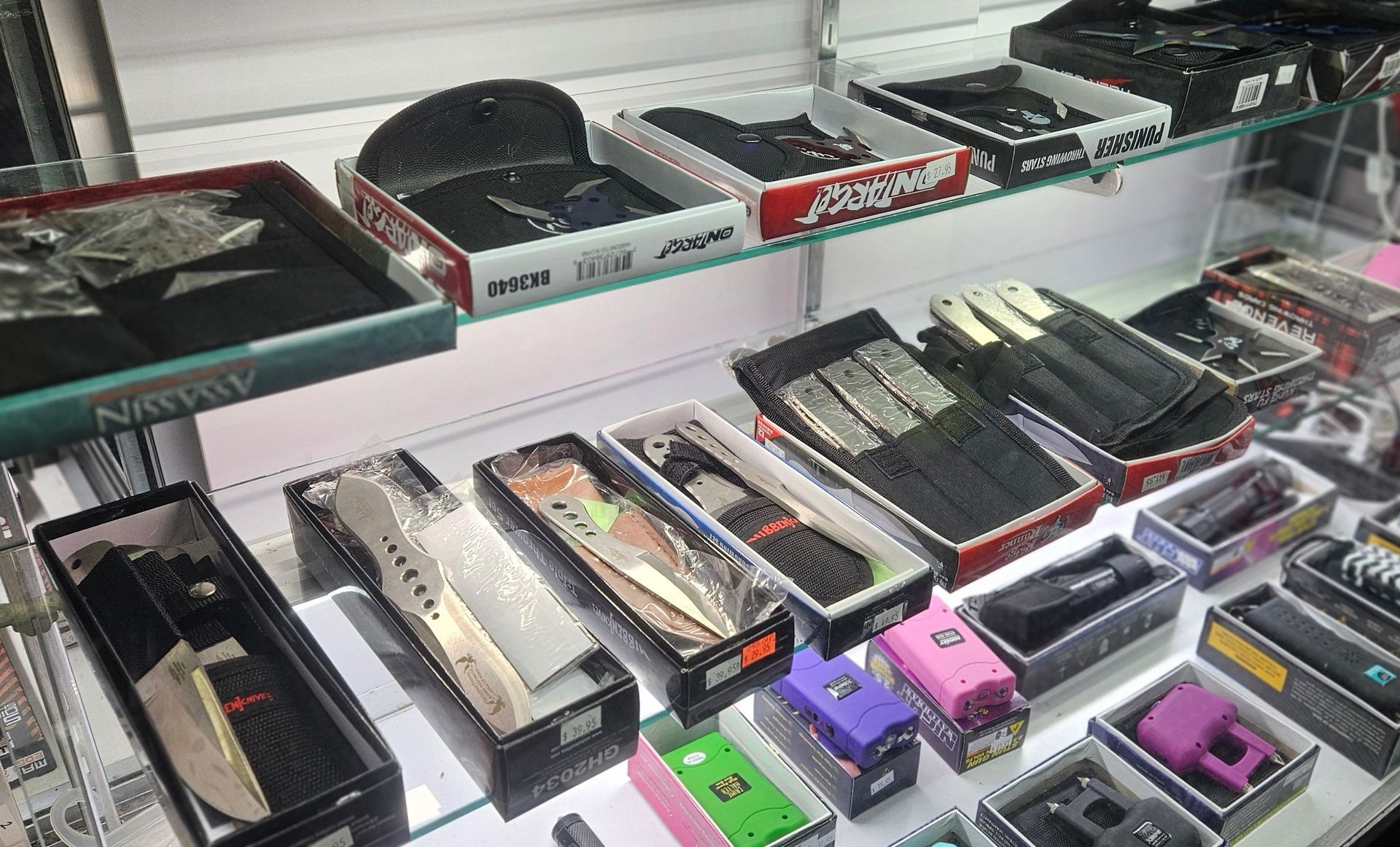 A case with a variety of throwing knives and stars on a shelf above several different tasers.