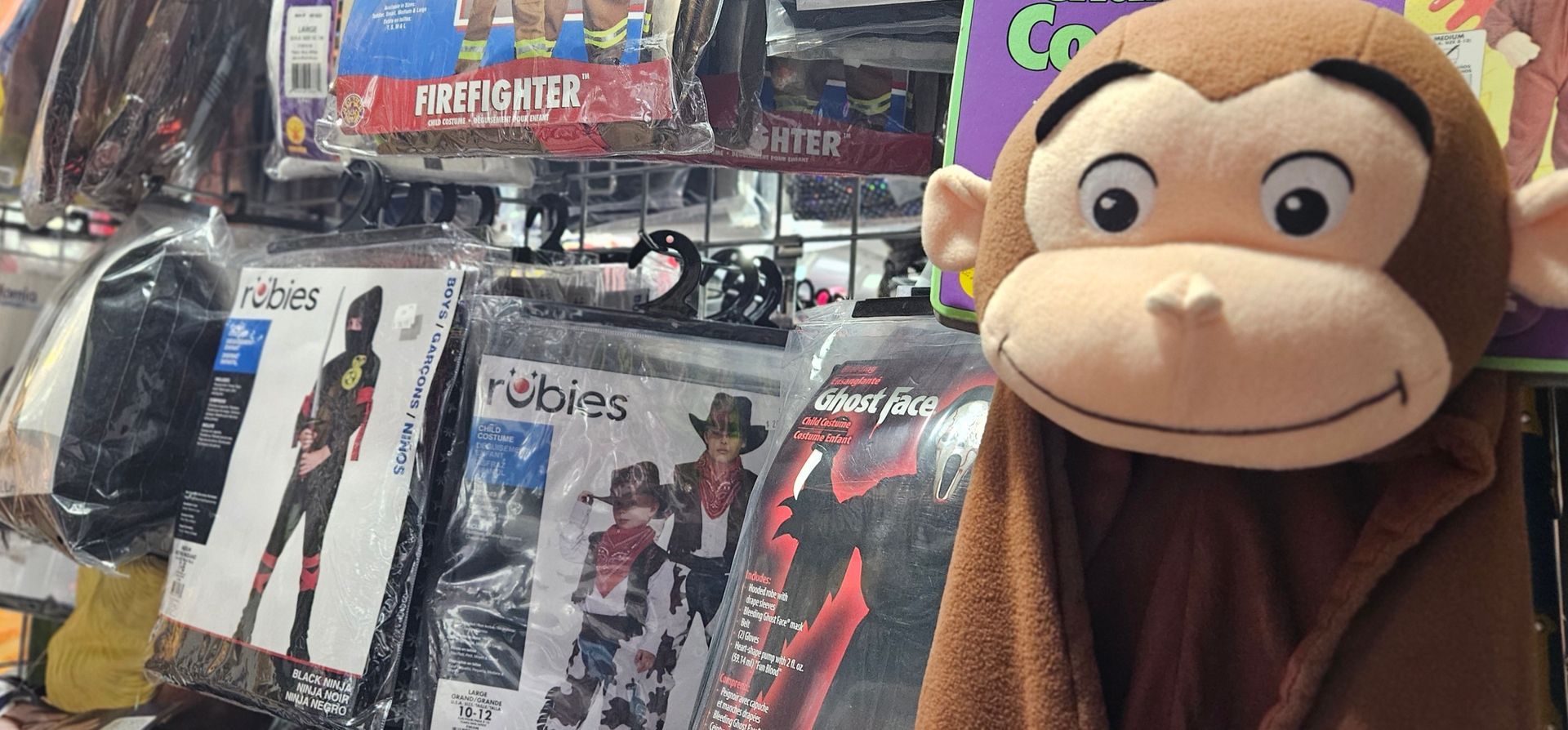 A monkey costume peers at the camera from in front of a display of various kids costumes by Rubies.