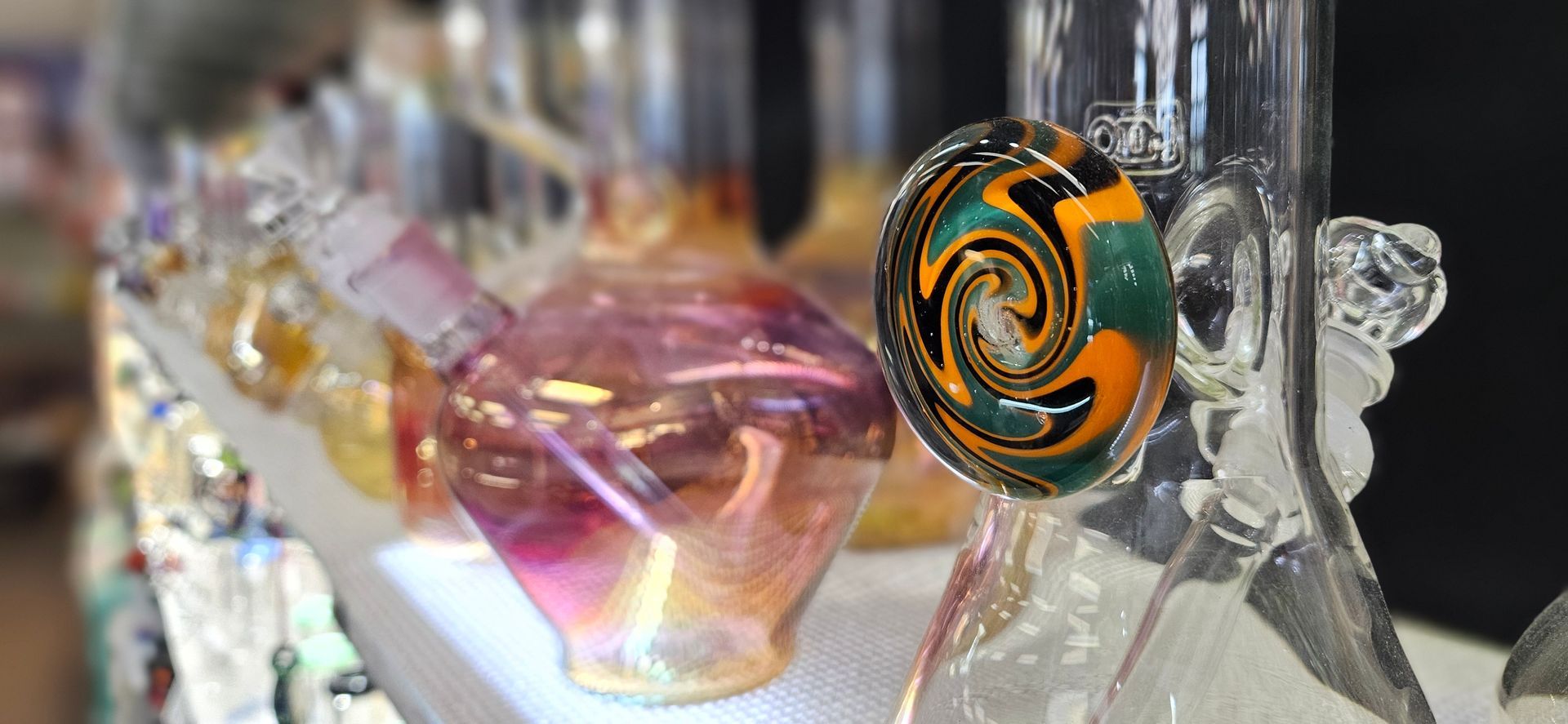A large and intricate wigwag marble is featured in focus while a shelf of glass bongs recedes into the background.