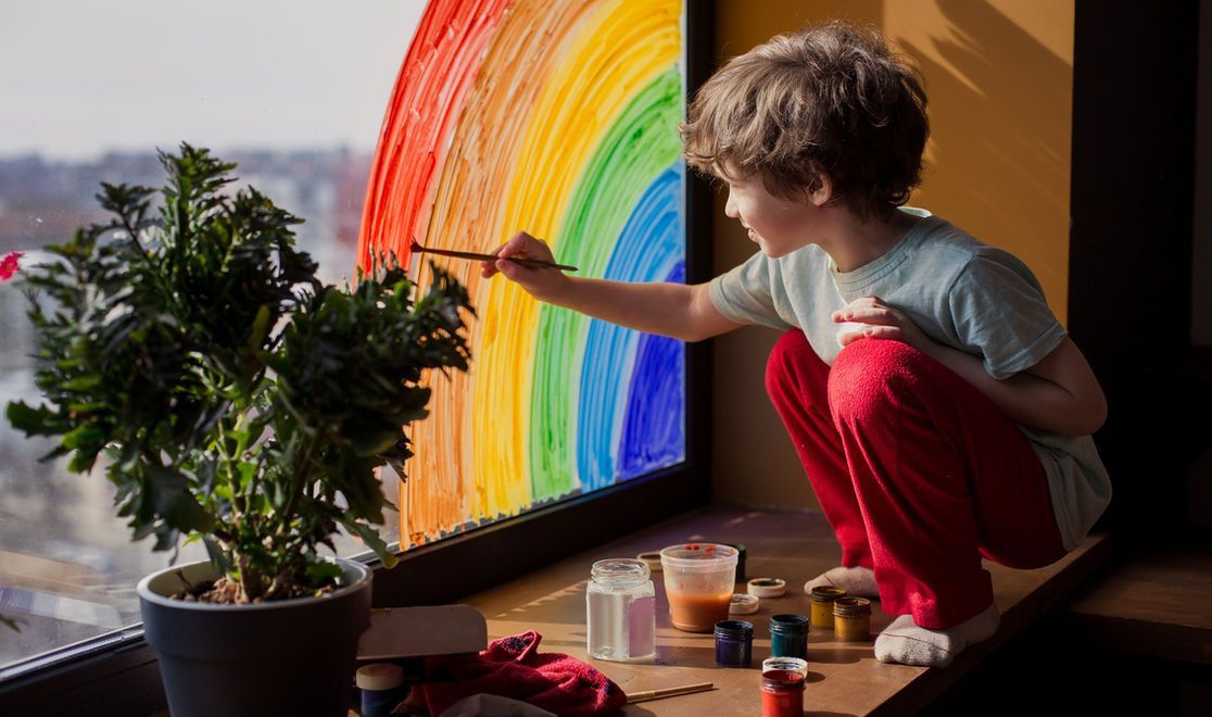 Boy painting a colorful rainbow on a window, squatting indoors with paint supplies nearby.