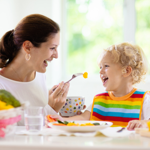 Woman feeding toddler with a fork, both laughing at a table. Brightly lit room.