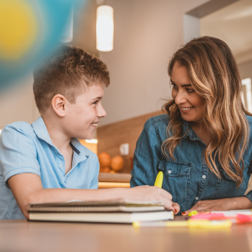 A smiling person and child look at each other while working on a table with school supplies in the home.