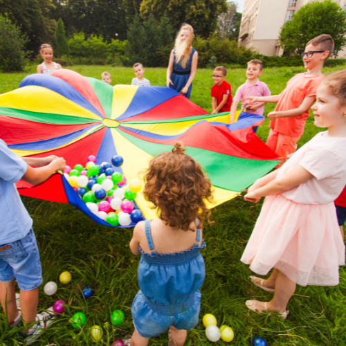 Children playing with a rainbow parachute on a grassy field, holding edges and balls inside.