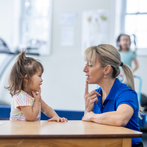 Speech therapist working with a young child, finger to lips, in a therapy room.