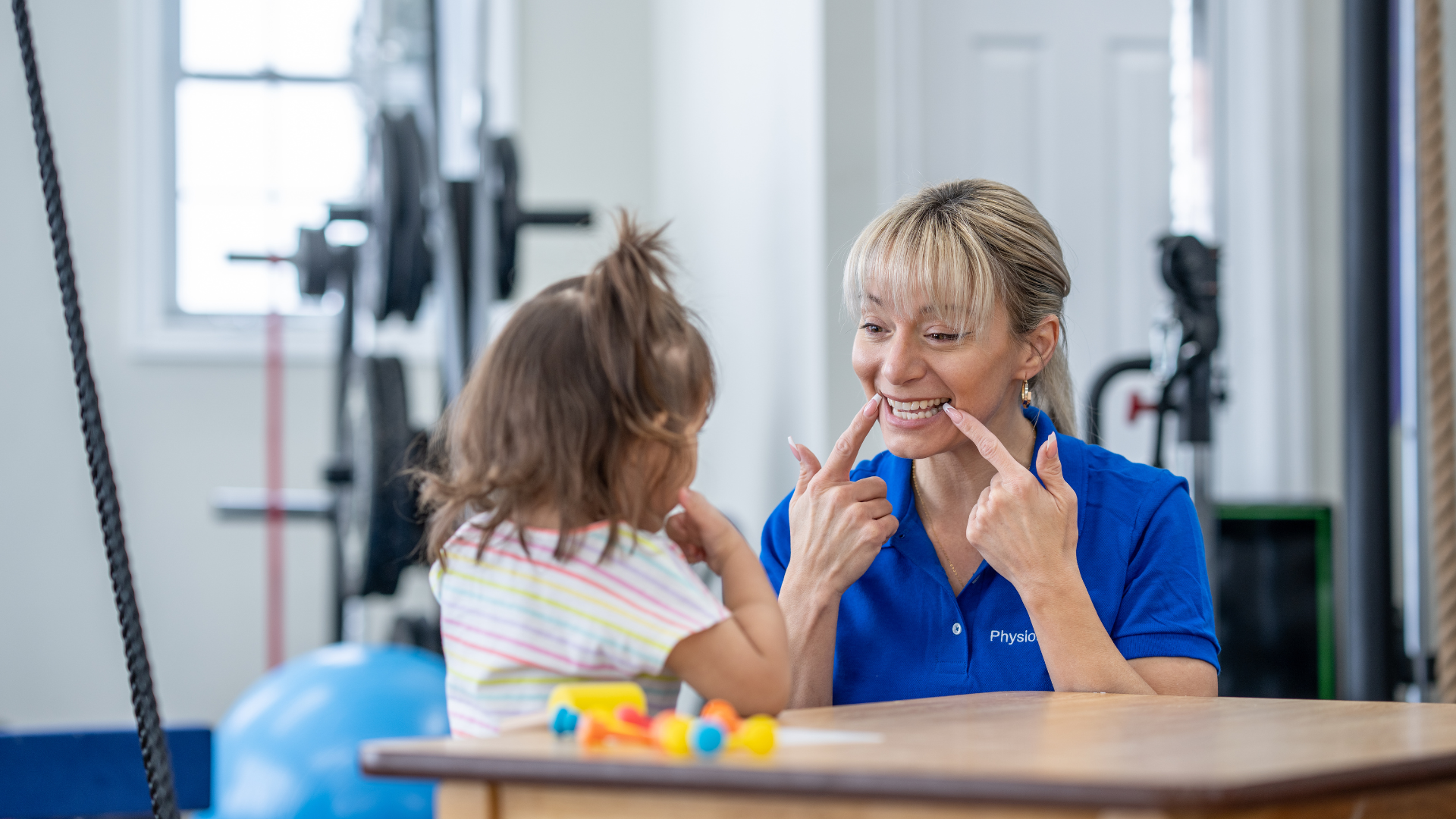Woman and child at table, woman pointing at her mouth. Bright room, fitness equipment in background.