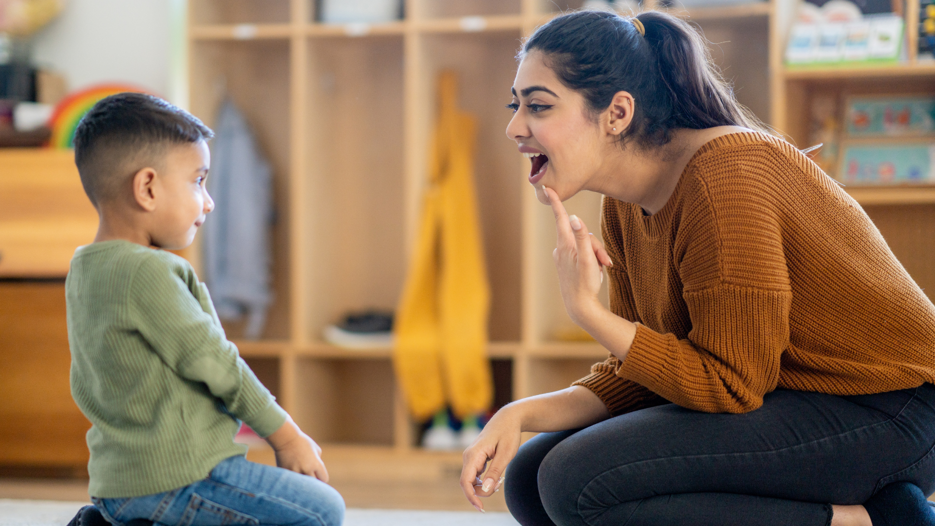 Woman kneels, demonstrating speech sound for child. Classroom setting, neutral colors.