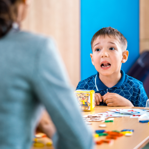 Child in blue shirt with open mouth at a table, looking towards a person out of frame.