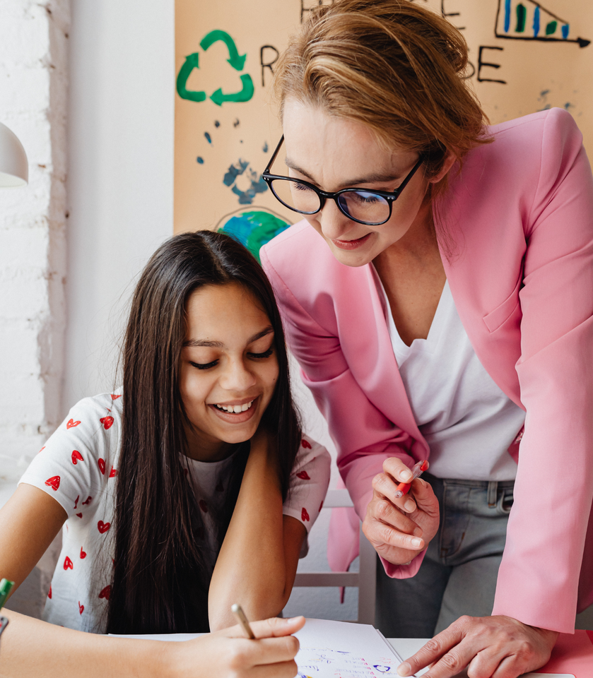 Woman in pink jacket assists smiling girl with schoolwork at a desk.