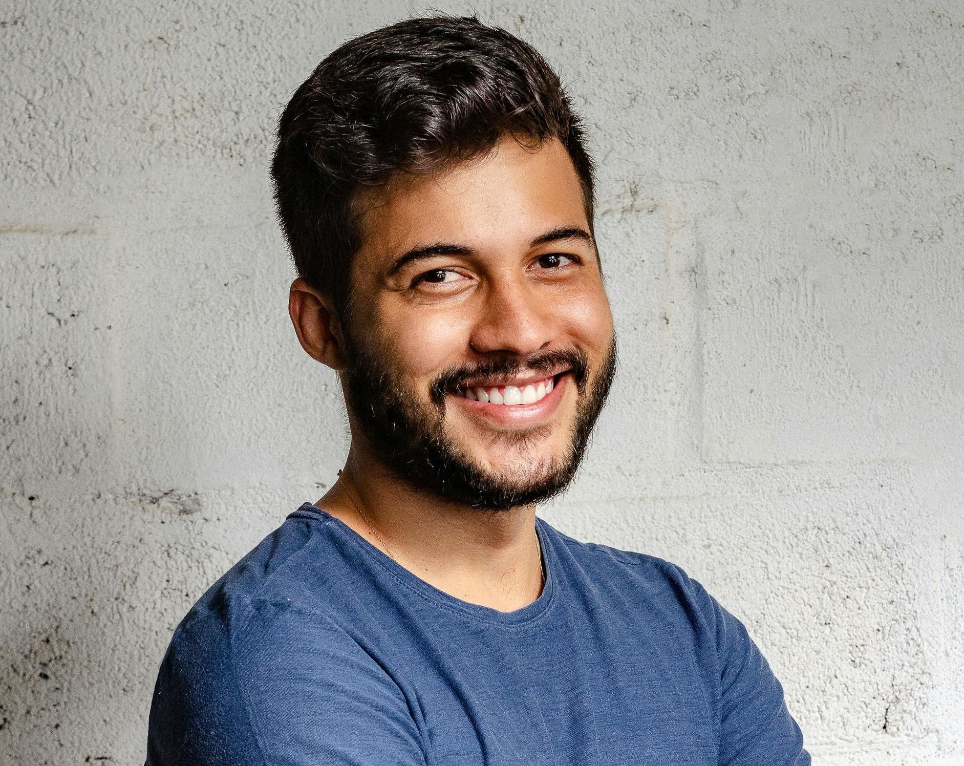 Man with a beard smiling against a textured, light gray wall, wearing a blue shirt.