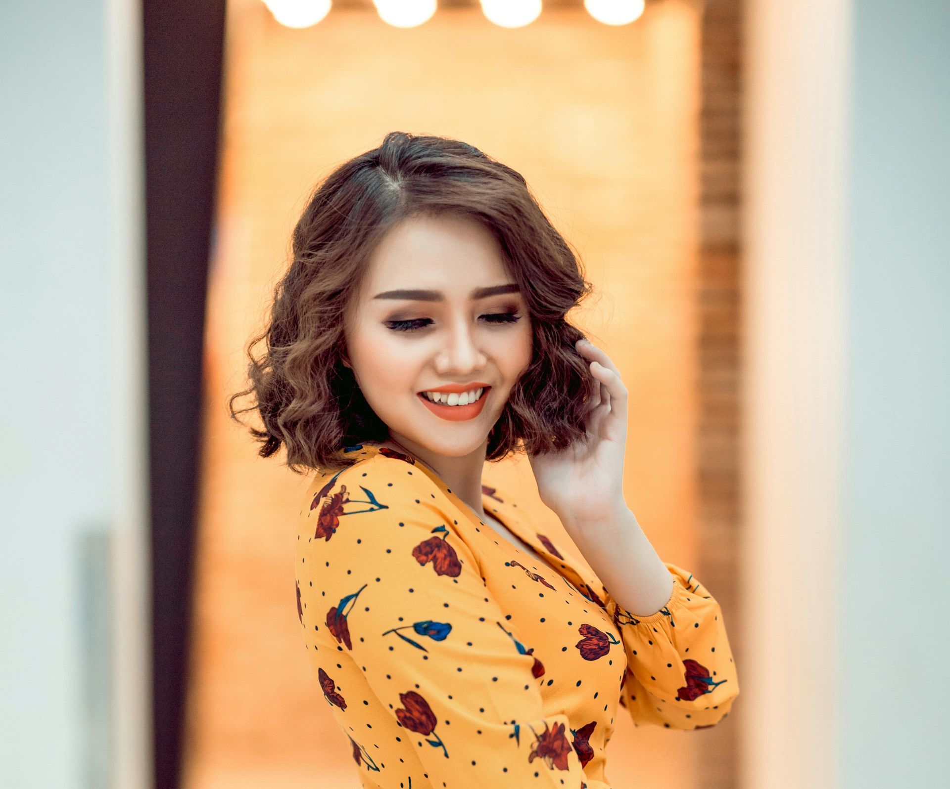 Woman with curly brown hair smiles while touching her hair; she's wearing a yellow floral top.