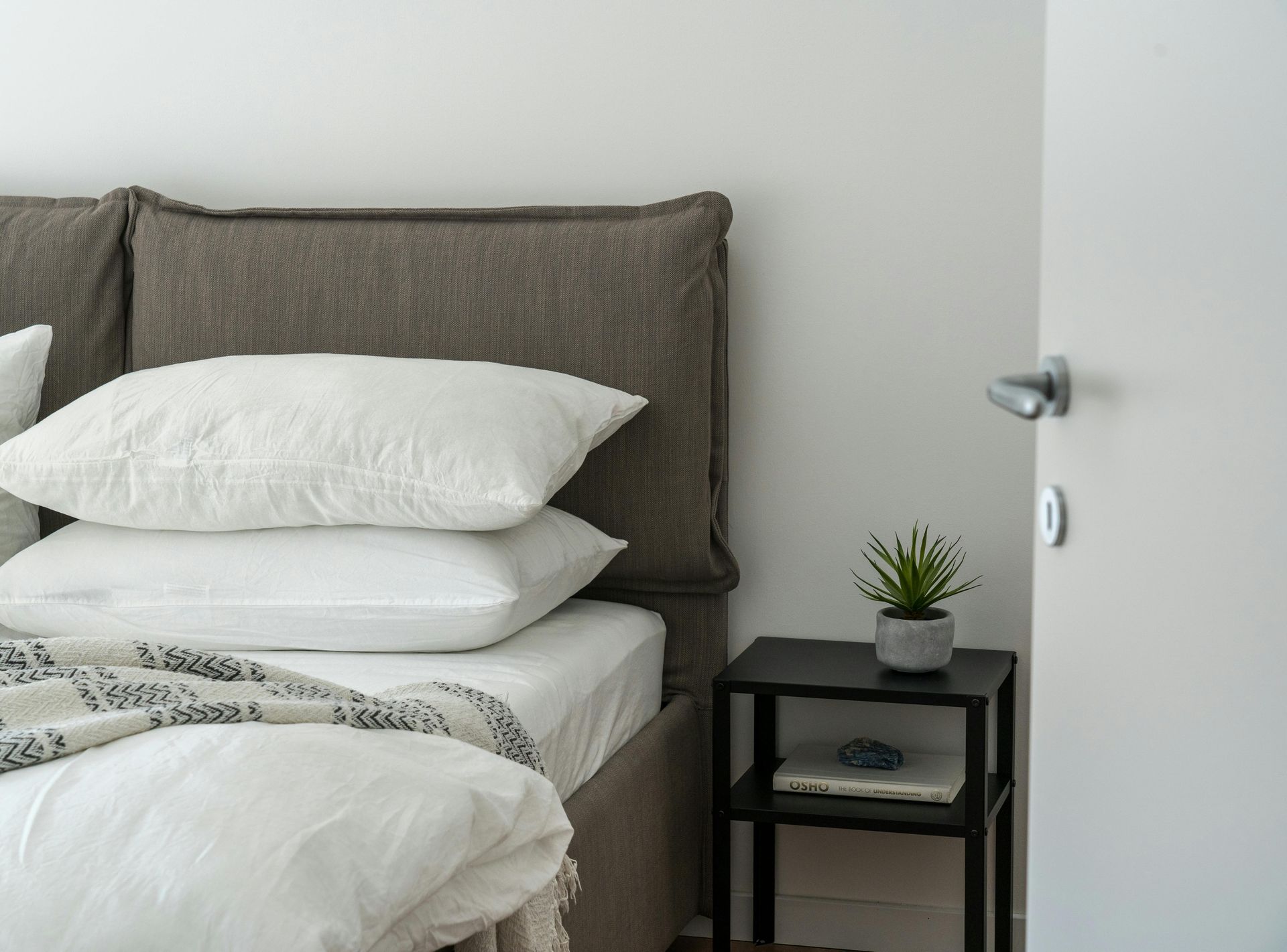 A bed with white sheets and pillows and a nightstand with a plant on it.