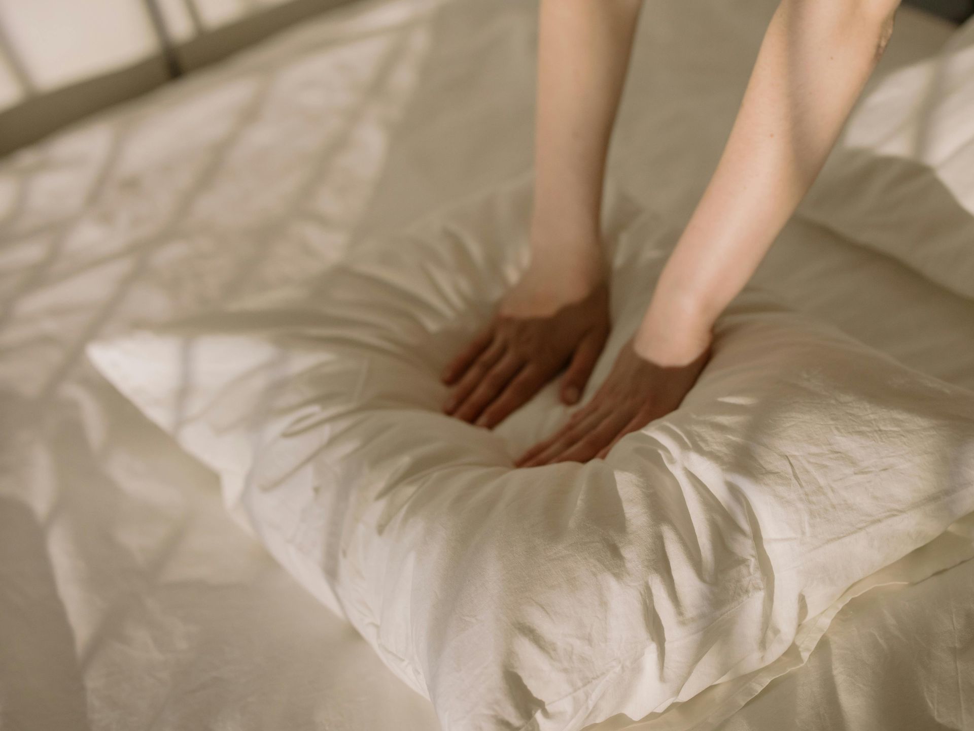 A person is putting their hands on a white pillow on a bed.