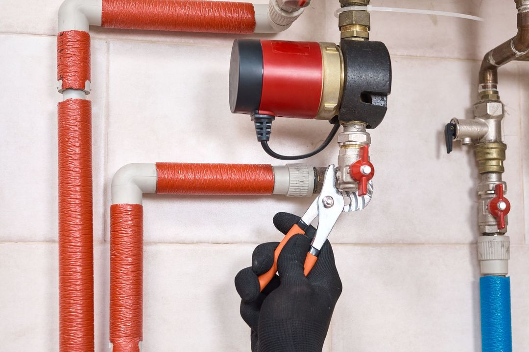 A person's gloved hand using pliers on a valve connected to water pipes and a red pump.