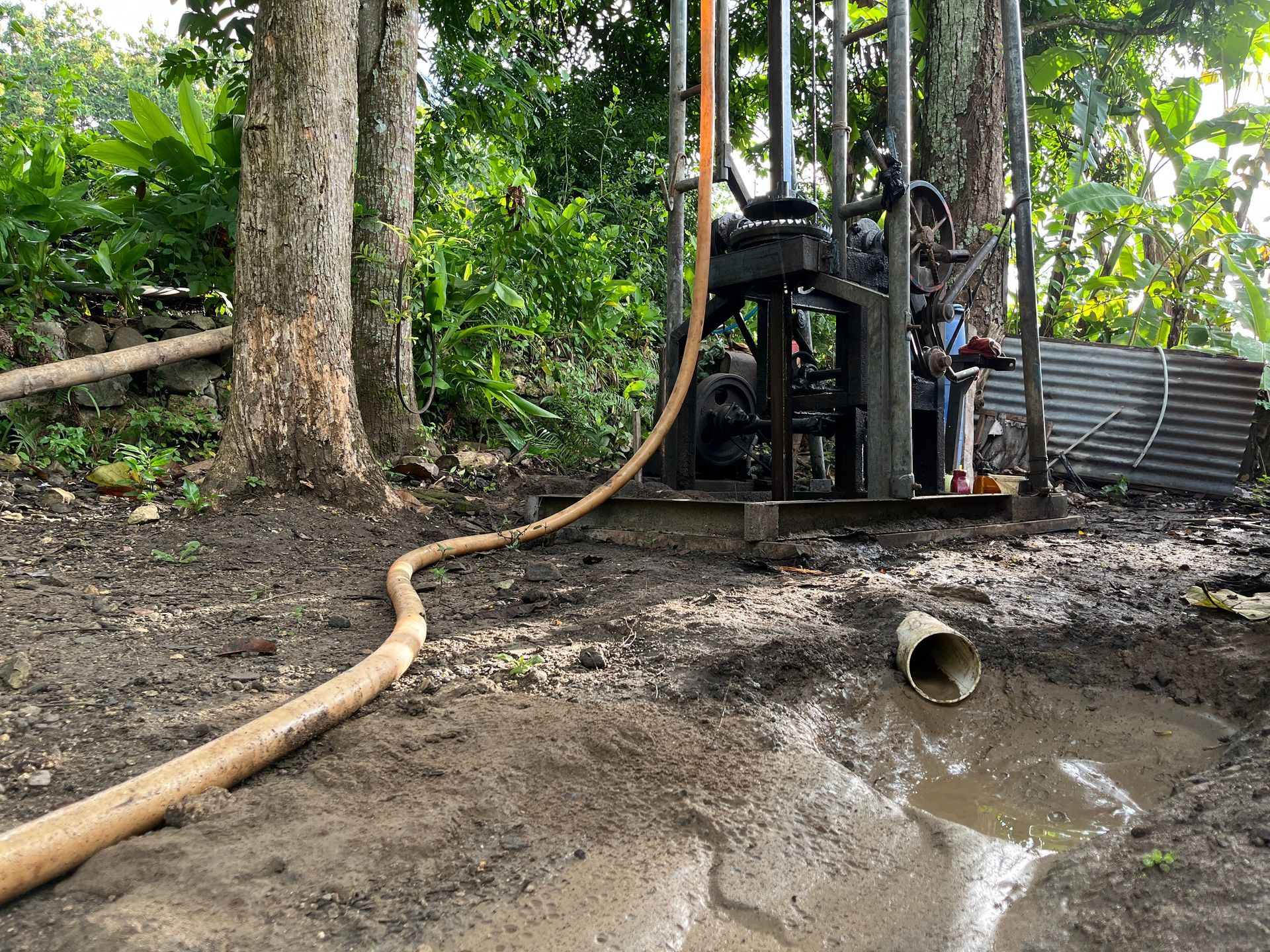 A drill rig operates in a muddy, wooded area, with a long hose extending toward a drainage pipe in a shallow pool. A drill rig operates in a muddy, wooded area, with a long hose extending toward a drainage pipe in a shallow pool.