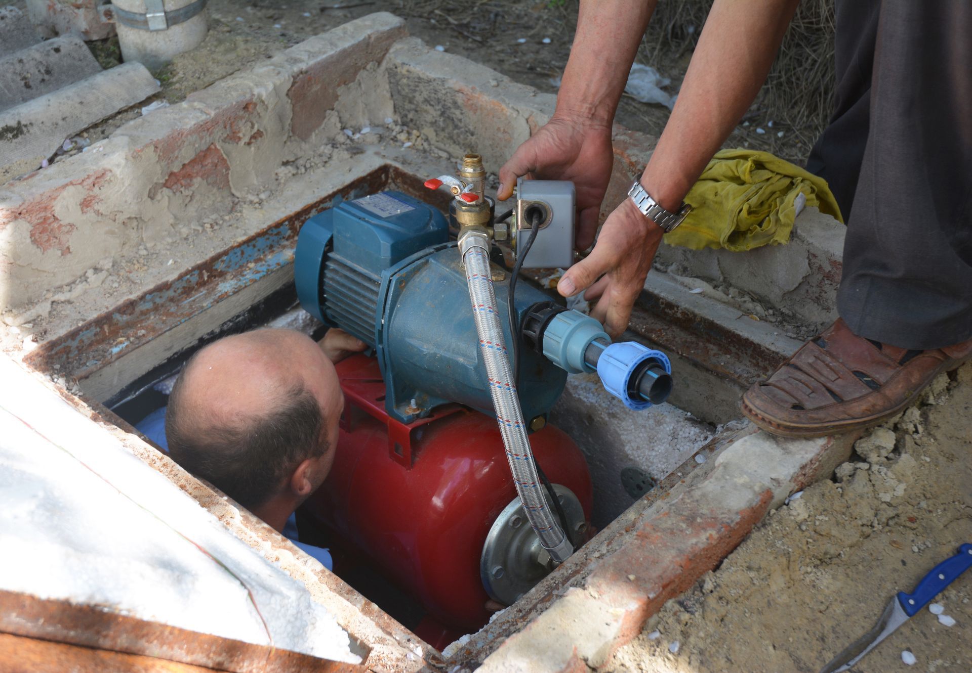 Two men installing a water pump in a brick-lined pit. One man leans in, the other holds wiring.