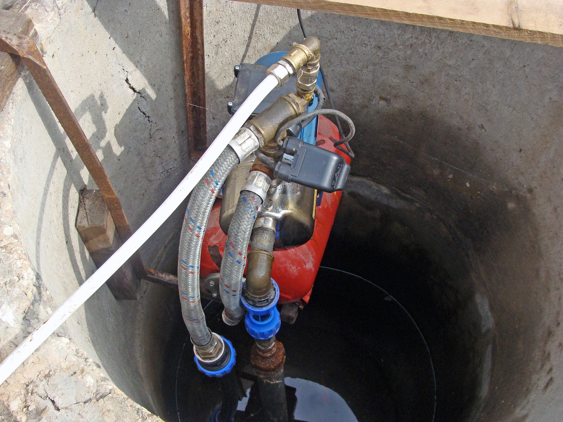 A red water pump with connected pipes inside a concrete well, partially submerged in water.