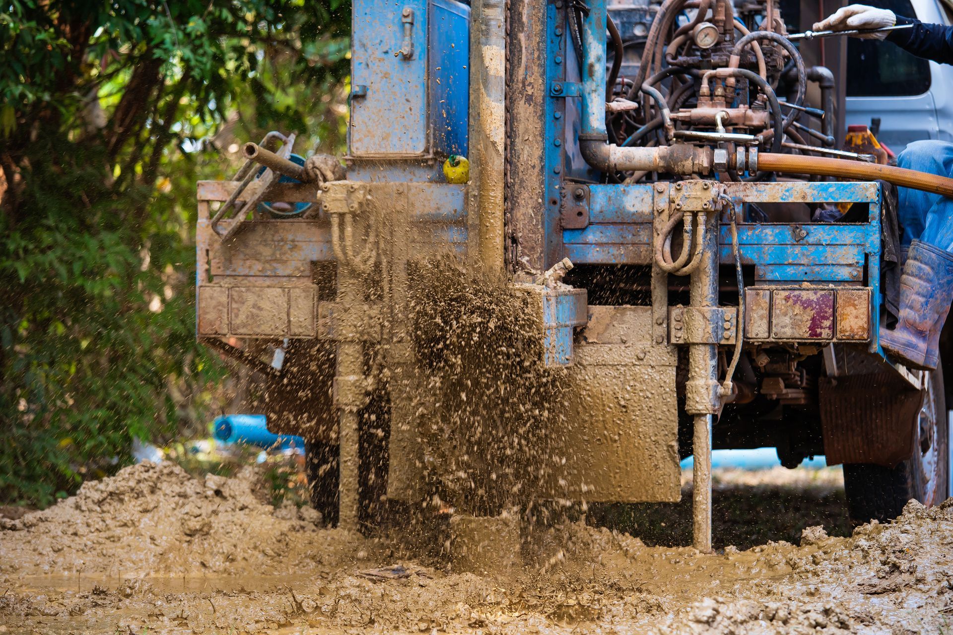 Drilling rig, blue and rust-colored, ejecting mud onto the ground during well construction, outdoors.