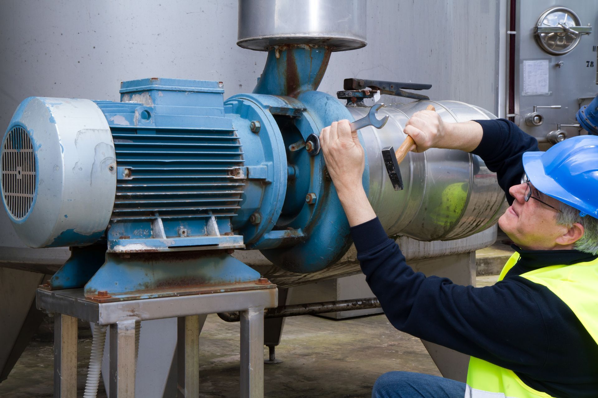 Mechanic working on a blue industrial pump with a wrench, wearing a blue hard hat and safety vest.