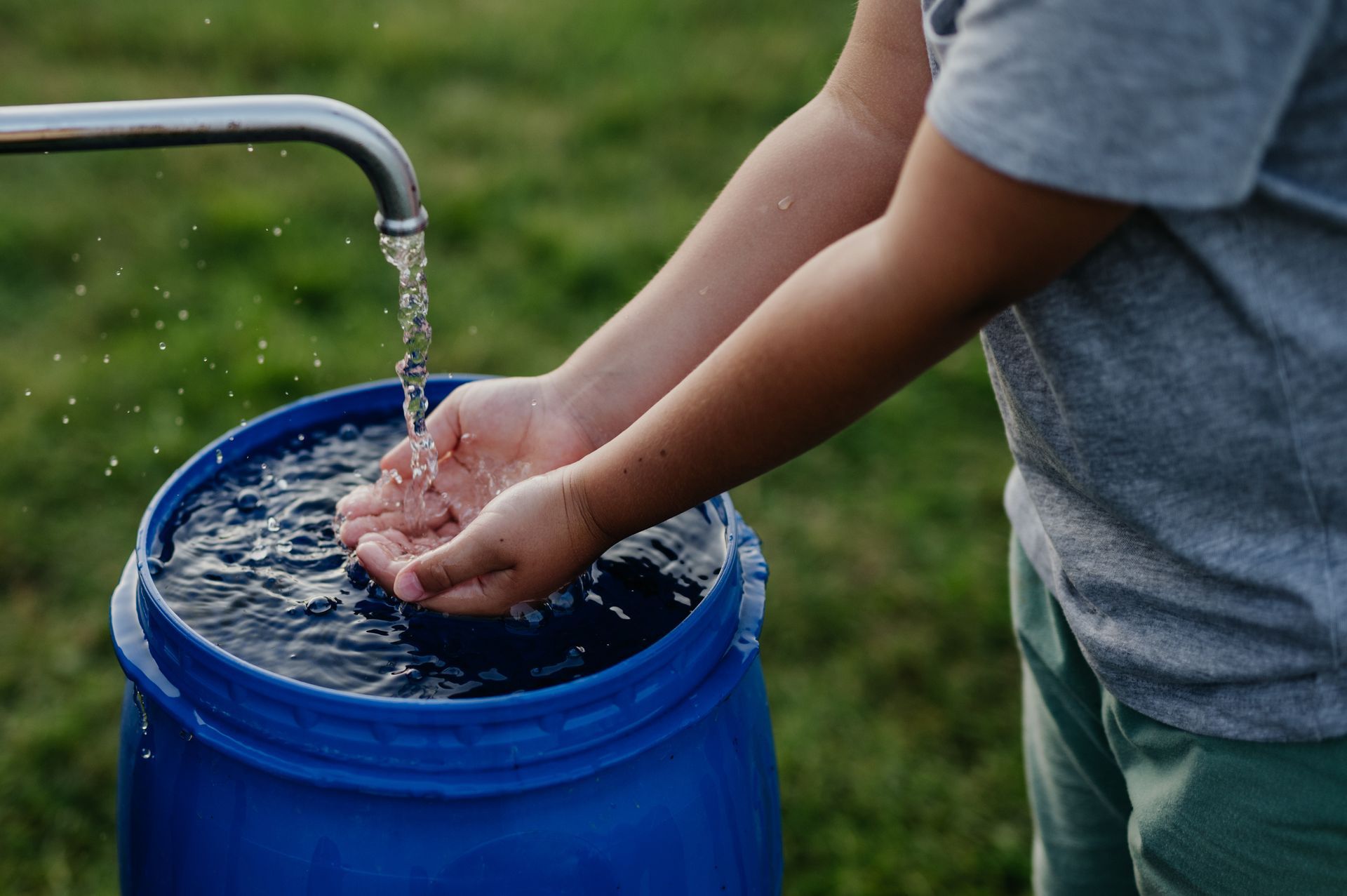 Child washing hands under a faucet, water flowing into a blue barrel. Outdoors, green grass.