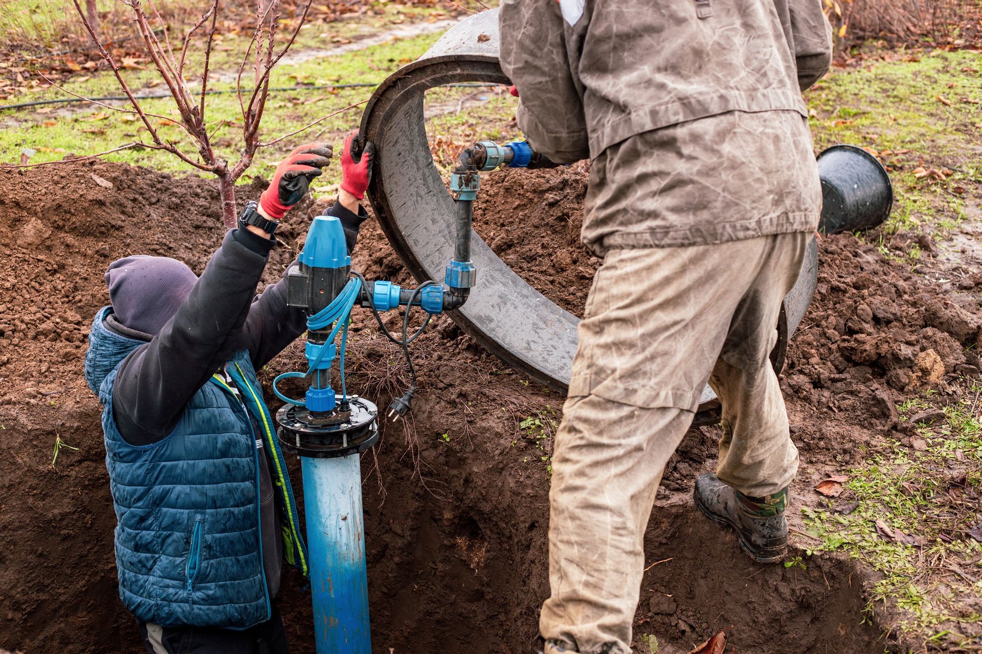 Two workers installing a water well, one in hole, other holding a concrete ring, outdoors, muddy ground.