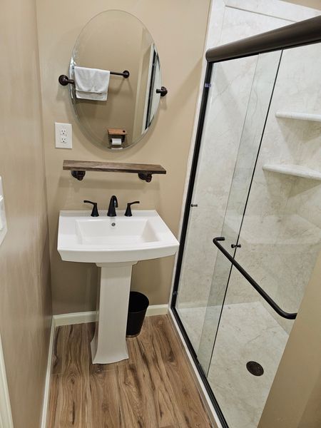 Small bathroom with a pedestal sink, oval mirror, and glass shower. Beige walls, dark brown accents, and wood-look flooring.
