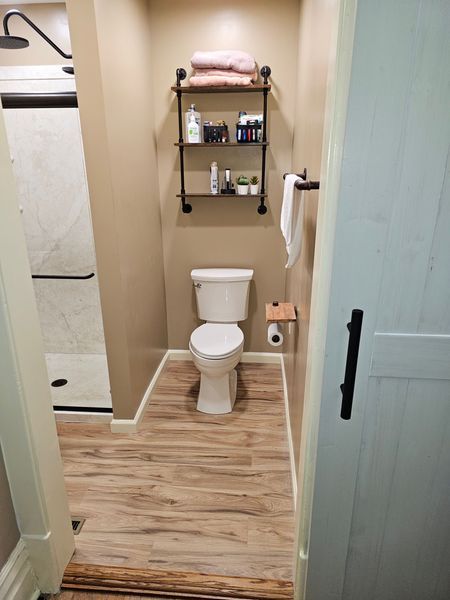 Bathroom with toilet, shower, and shelving unit. The room has a light wood-look floor and beige walls.