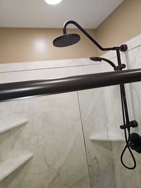 Dark bronze showerhead and handheld sprayer against a marble-look shower wall. A long bar runs across the shower.