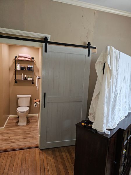 A light gray barn door slides open to reveal a small bathroom with a toilet, shelf, and towel rack. Brown floors and walls.