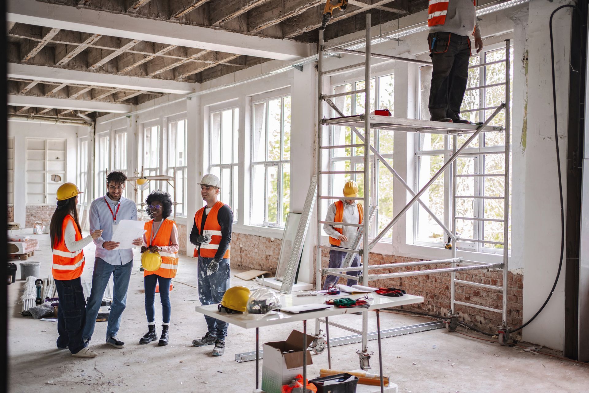 Construction workers in a building with scaffolding. Some are looking at plans, others working.  White walls, large windows.