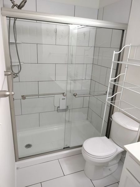 White tiled bathroom with a glass shower, toilet, and a shelf unit to the right.
