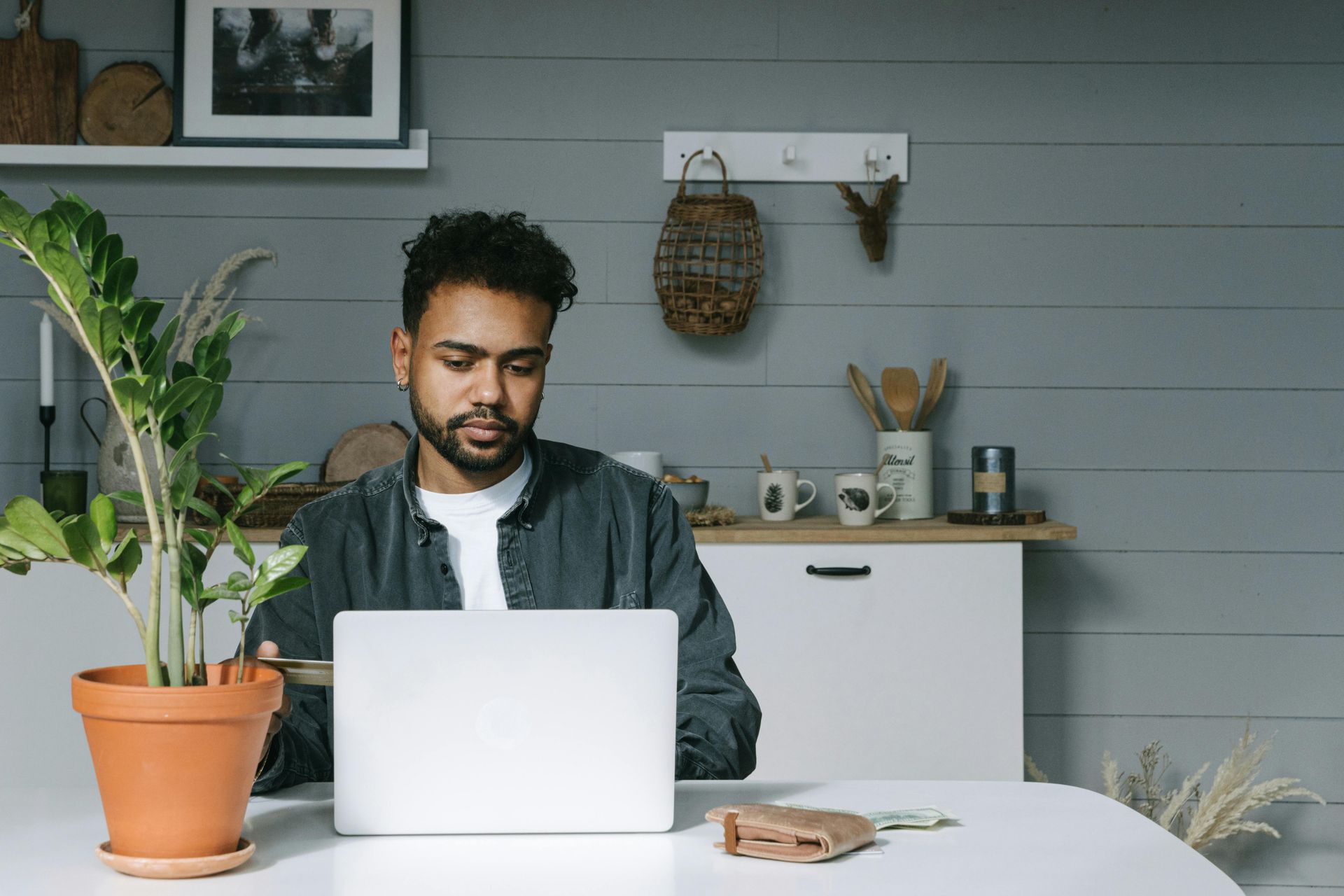 Man working on a laptop at a white table in a kitchen with a plant, against a gray wall.