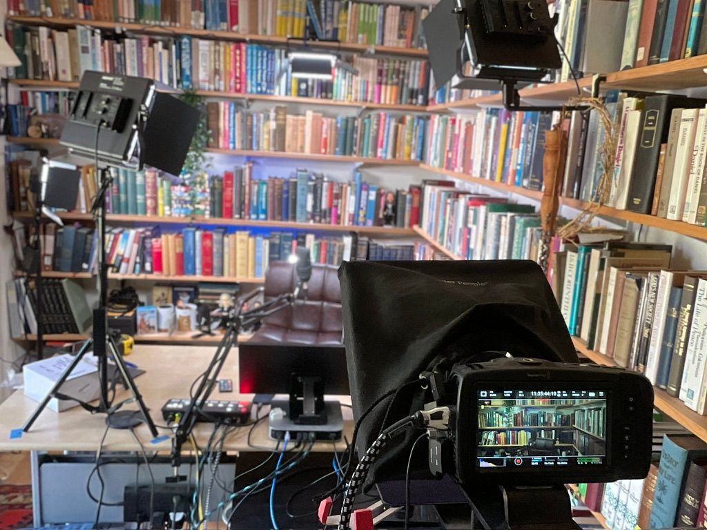 A cluttered home office with a camera set up for filming, surrounded by bookshelves and bright lights.