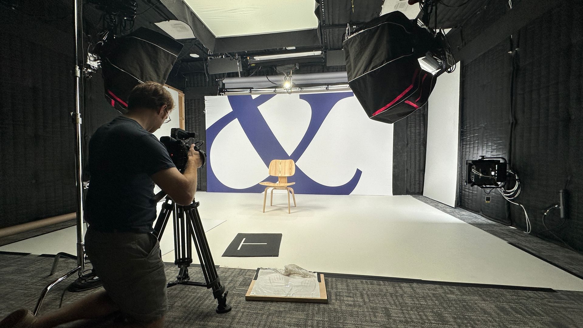 A man is in a photo studio with a chair and big backdrop with an "&" sign, setting up a video.