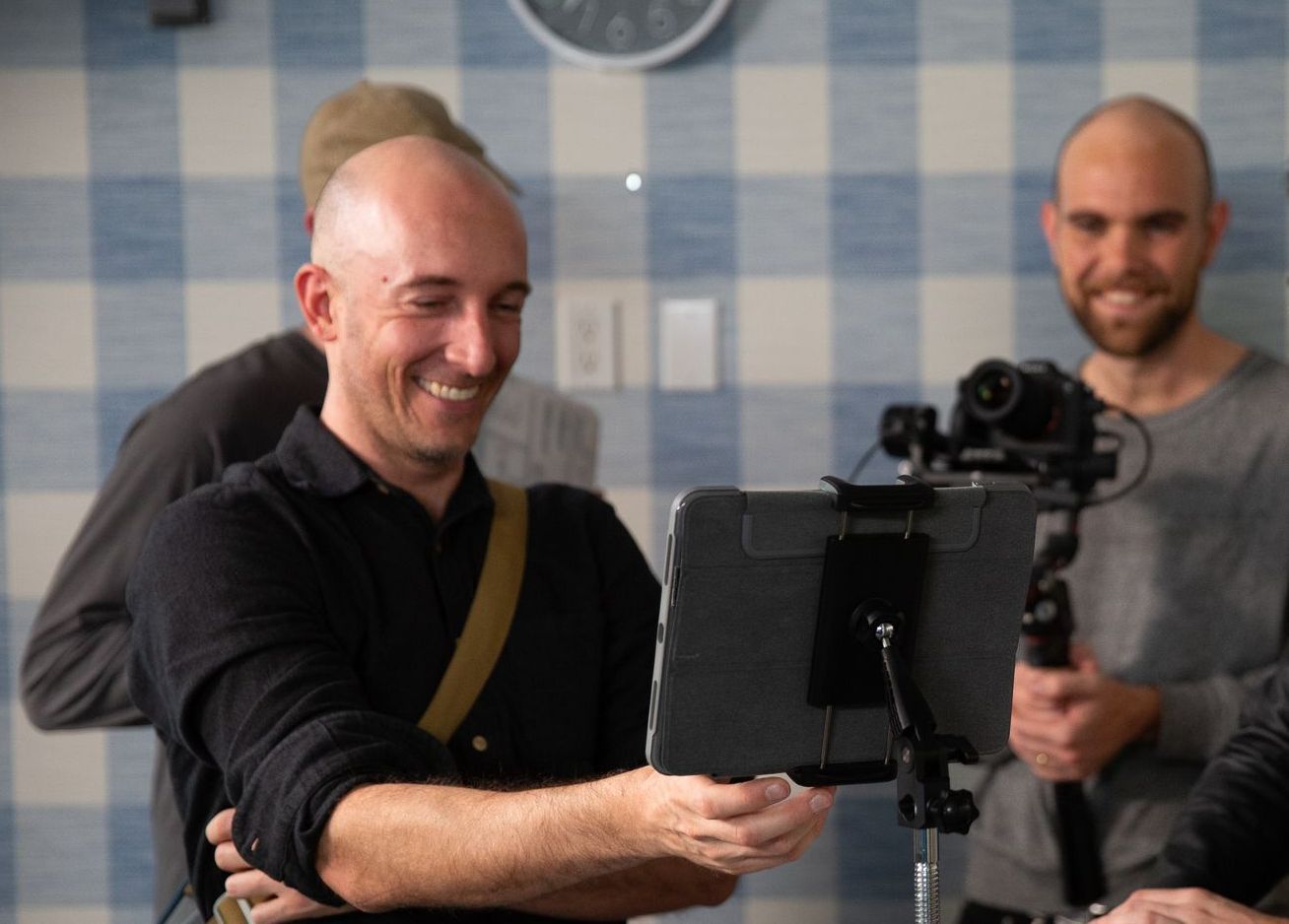 A man is smiling while holding a tablet in front of a camera.