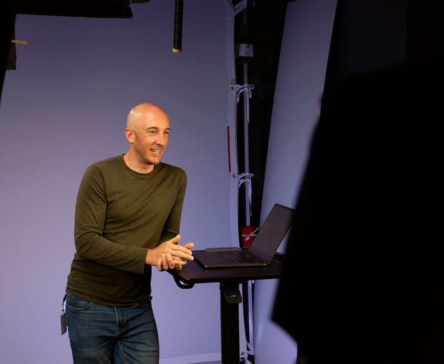 Bald man in green shirt at a laptop in a studio, smiling and gesturing.