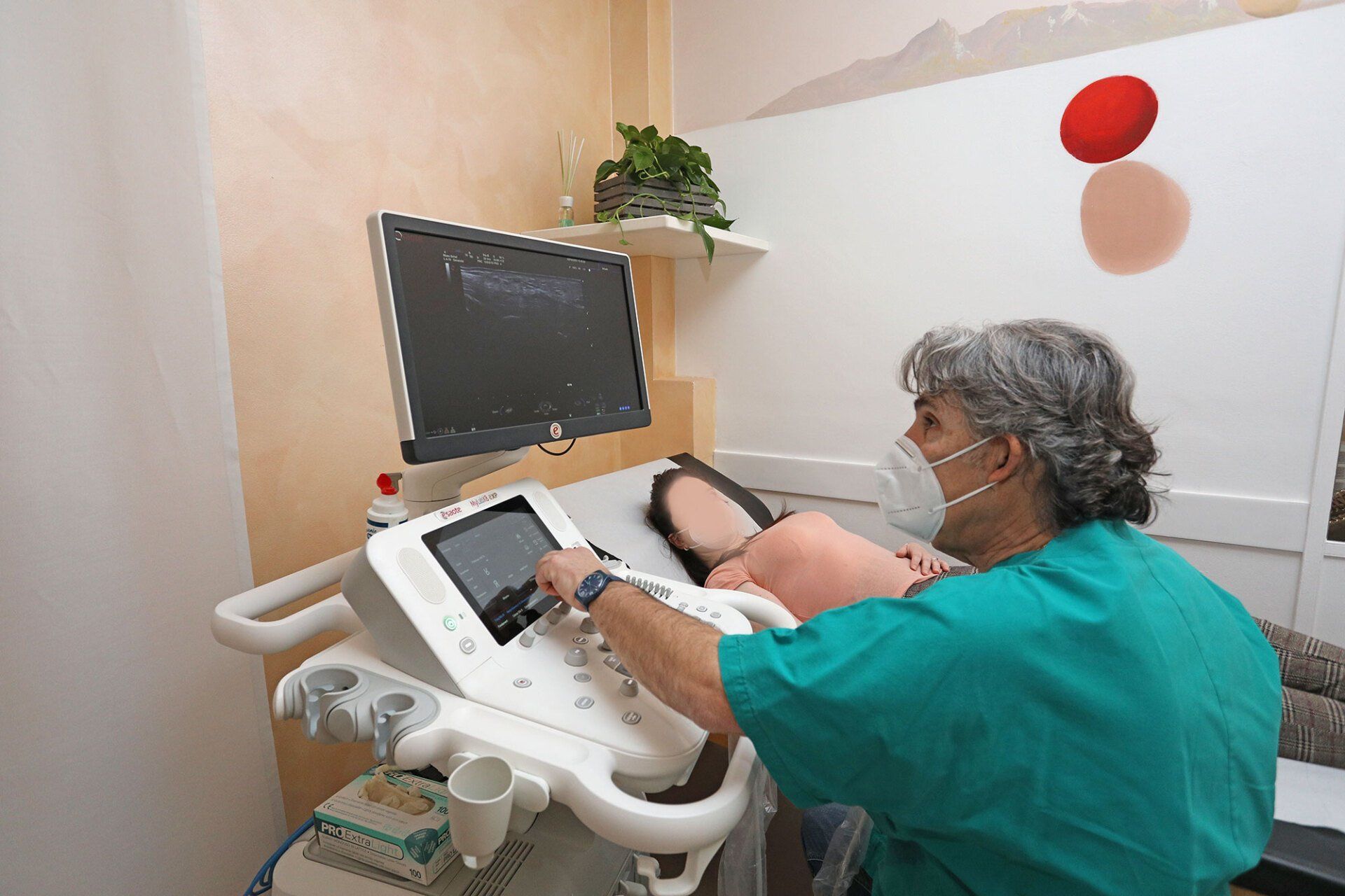 Un operatore sanitario con indosso una casacca verde utilizza un apparecchio a ultrasuoni per esaminare un paziente in una stanza clinica.