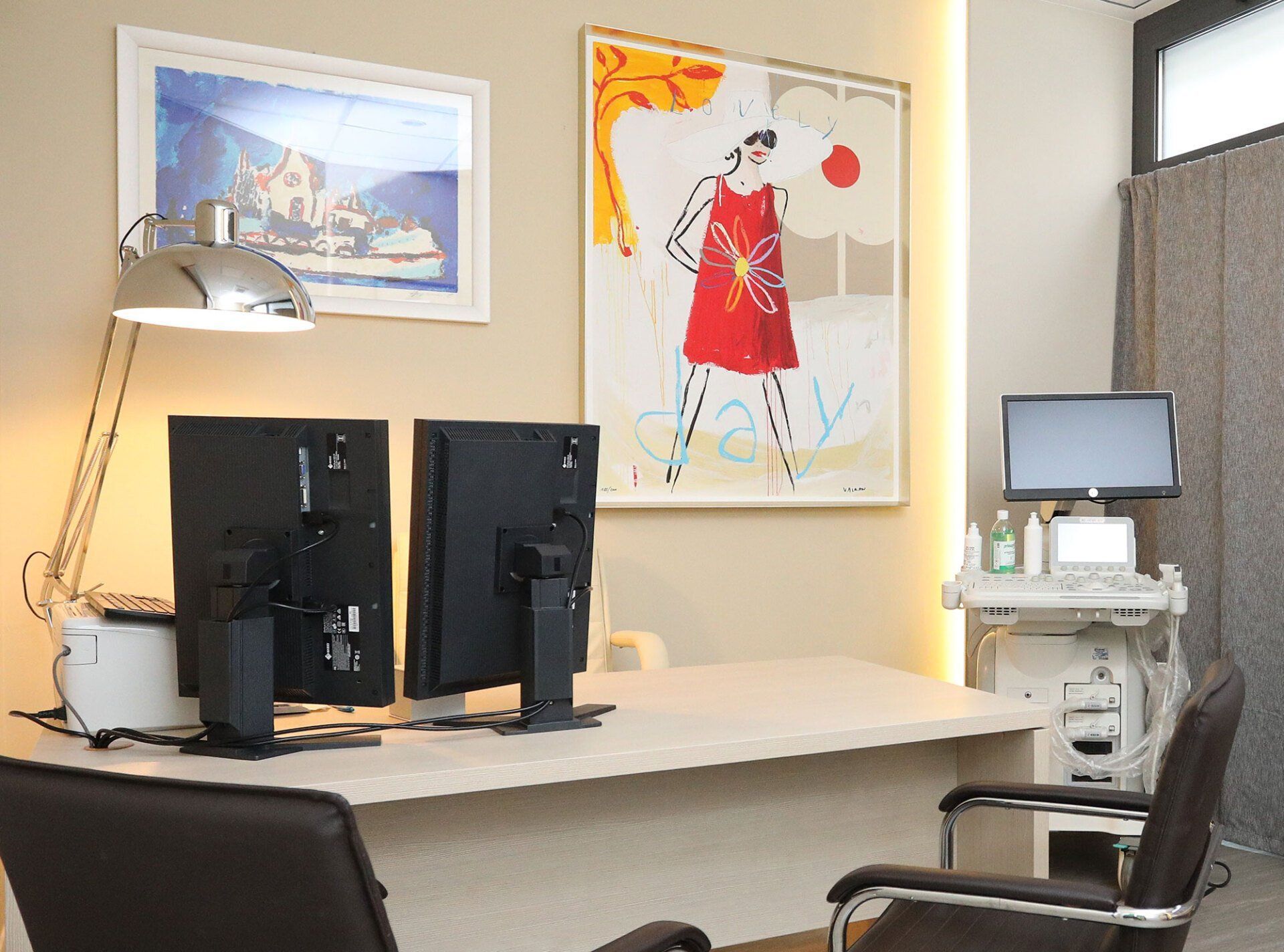 Uno studio medico con una scrivania, due monitor, un apparecchio per le visite mediche e opere d'arte alle pareti.