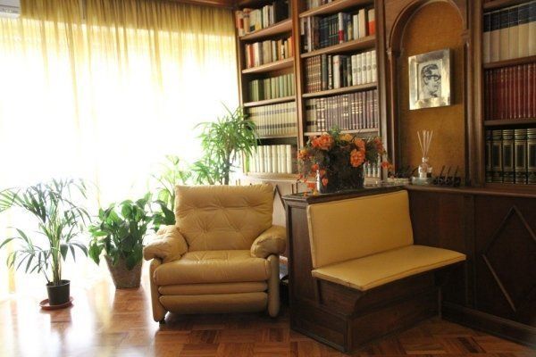 Una stanza adibita a biblioteca domestica con scaffali in legno, una poltrona beige, una panca abbinata e diverse piante in vaso.