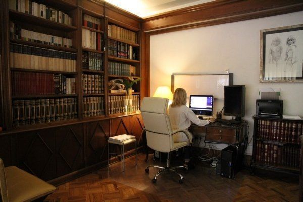 Una persona siede su una sedia da ufficio bianca a una scrivania con un computer in una biblioteca domestica piena di scaffali che vanno dal pavimento al soffitto.
